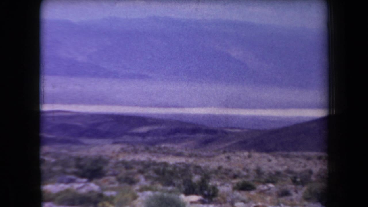 Wide desert view with brush, low hills, distant mountains, and pale horizon light.