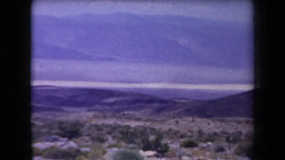 1958 Arizona home-movie frame with sparse desert vegetation, broad hills, distant mountains, and soft purple color fading.