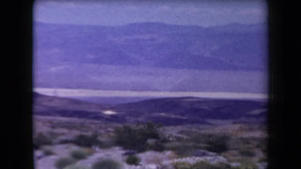 Arizona 1958 8mm still with desert shrubs in the foreground, dark rolling hills, and layered mountains under a faded sky.