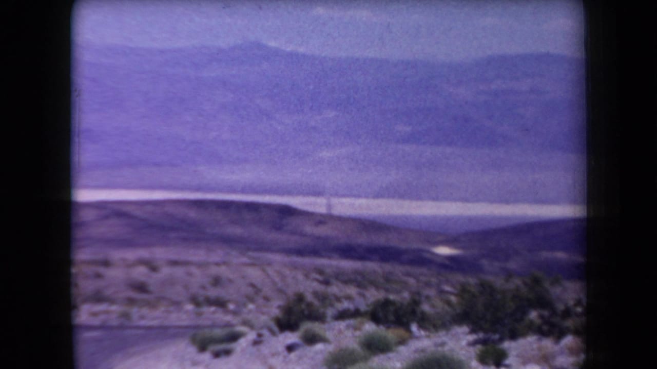 Wide desert landscape with shrubs, rolling hills, distant mountains, and a hazy sky.
