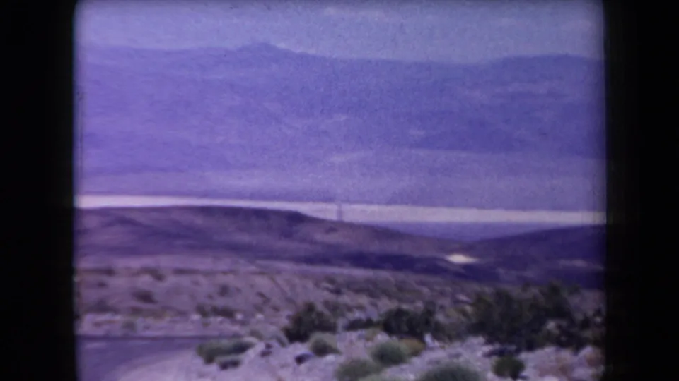 1958 Arizona 8mm frame showing a wide desert view with low shrubs, bare hills, distant mountains, and a purple-tinged sky.