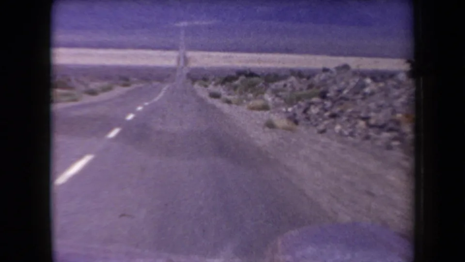 1958 Arizona road-trip frame from a moving vehicle, looking along an empty two-lane highway bordered by rocks and desert shrubs.
