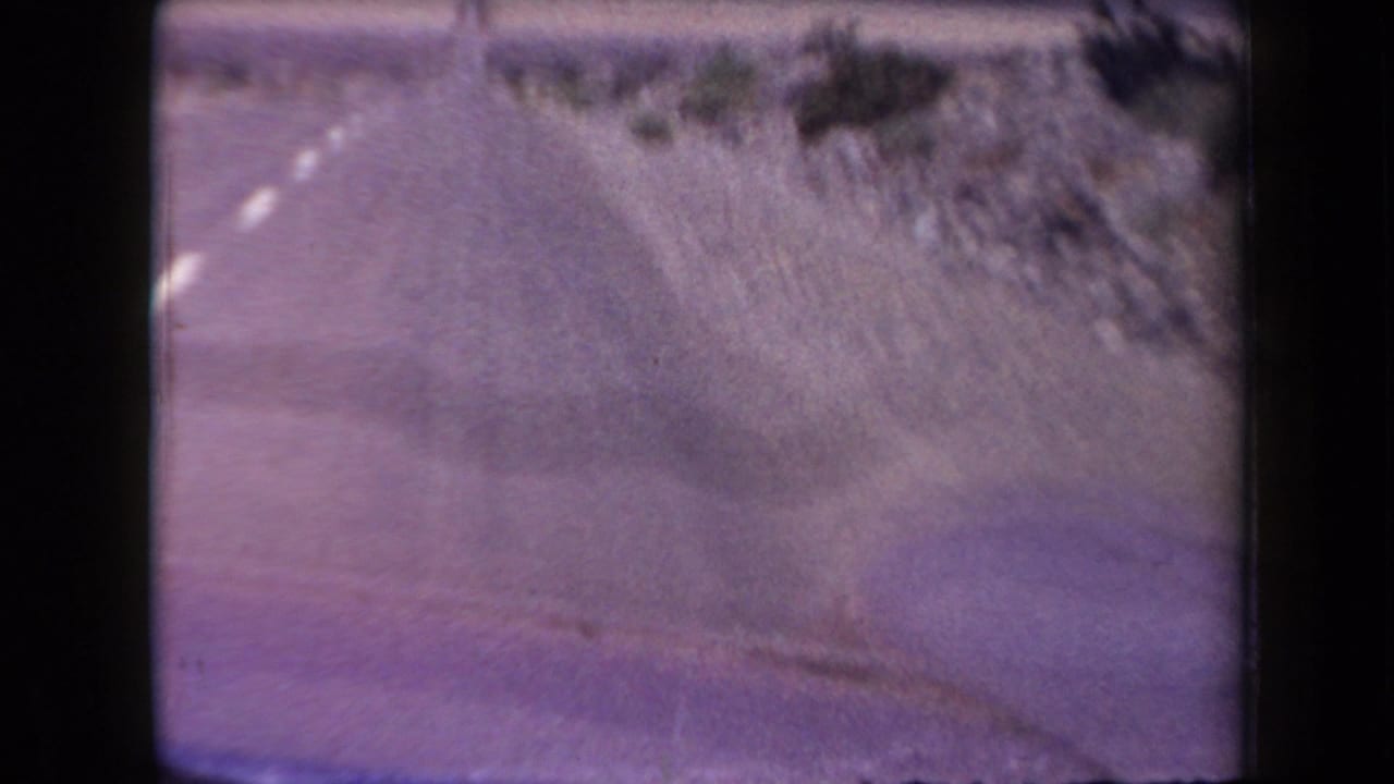 1958 Arizona 8mm frame showing a blurred forward view from a moving car on a two-lane desert highway with rocky roadside terrain.