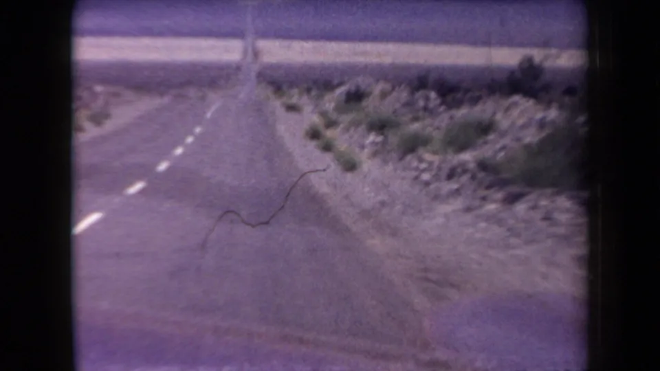 1958 Arizona 8mm frame from a moving car, looking along a two-lane desert road with gravel shoulder, scrub plants, and rocky ground.
