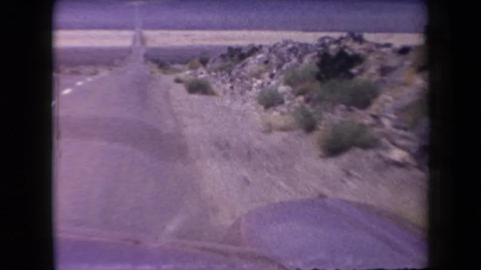 Forward view from a moving car on a 1958 Arizona desert highway, with the rounded hood edge visible below rocky roadside brush.