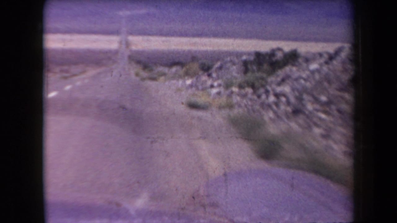 1958 Arizona 8mm road-trip frame showing a paved highway bordered by rocky desert ground and sparse shrubs.