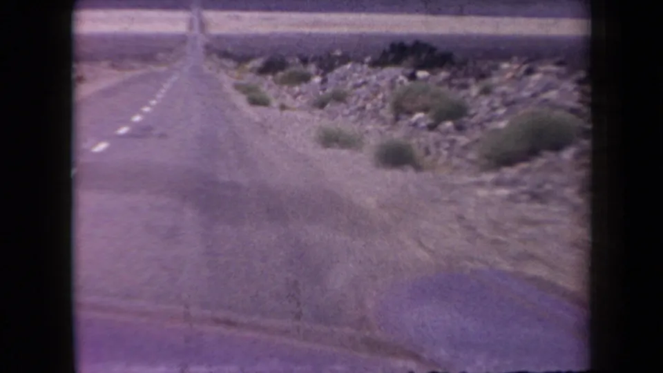 A soft 8mm frame shows the Arizona highway from a moving car, with desert rocks and scattered shrubs along the shoulder.