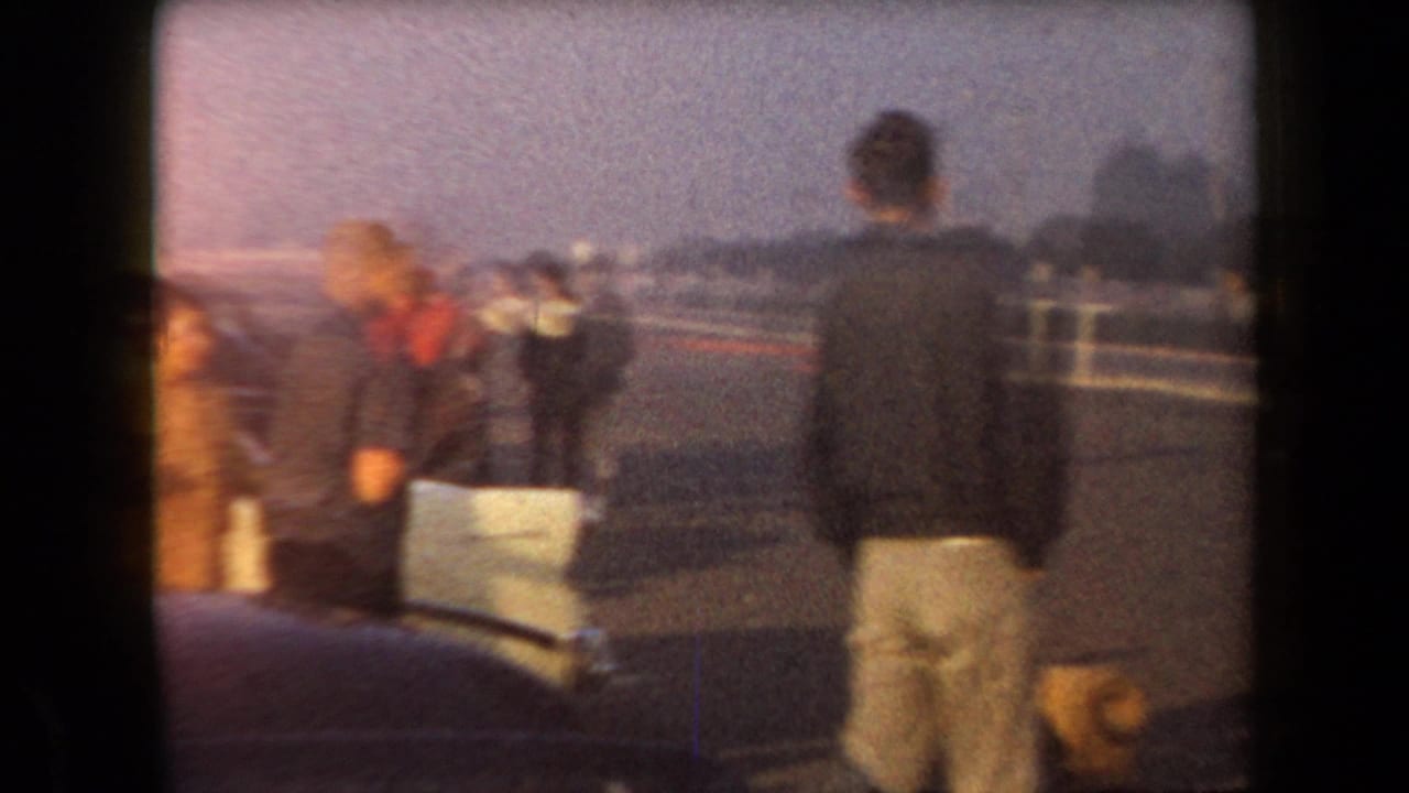 In 1958 Arizona home-movie footage, several people appear in a parking lot near cars under a dim evening sky.