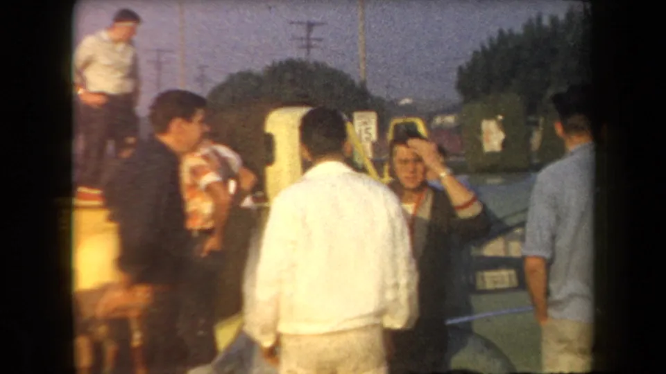 Young men in mid-century casual clothing gather beside a parked car in the Arizona parking-lot scene.