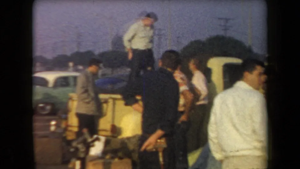 A handheld 8mm frame shows men in 1950s attire gathered around cars in the Arizona parking lot.