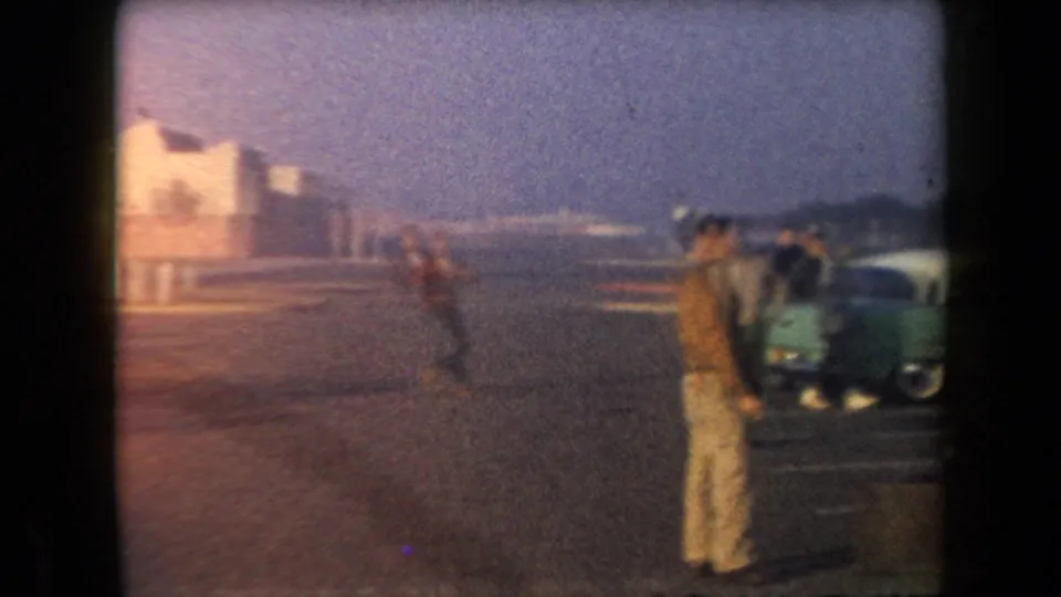 A wider 1958 Arizona frame shows people spread across a parking lot with parked cars and low buildings in the distance.