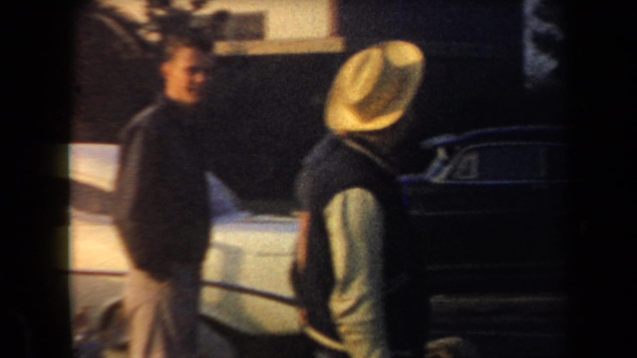 Two adults stand beside parked cars, one wearing a light hat near a dark sedan.