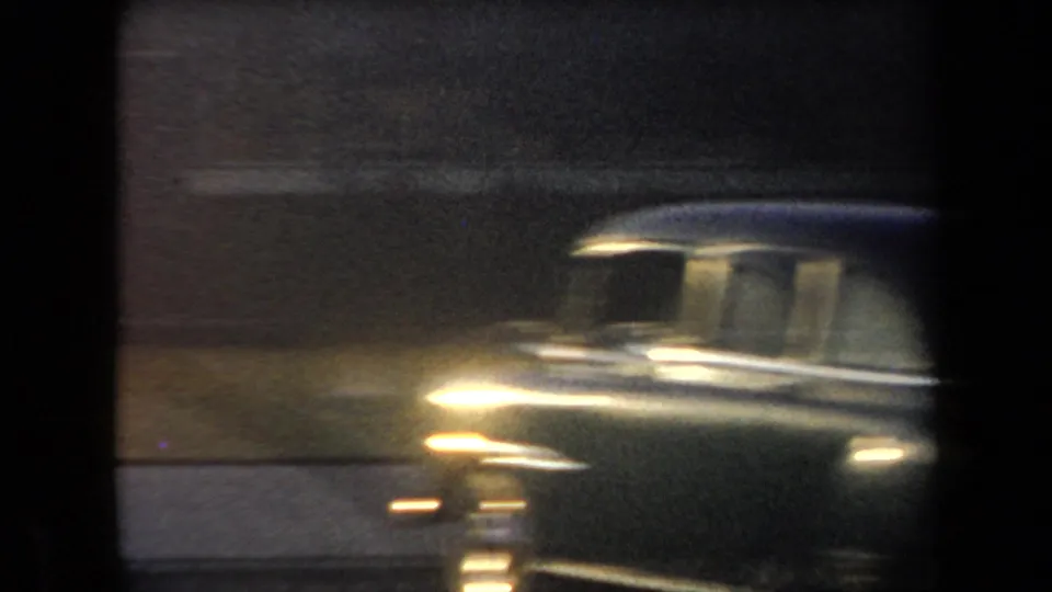 Soft 8mm frame of a dark mid-century car passing or parked near the roadside in dim exposure.