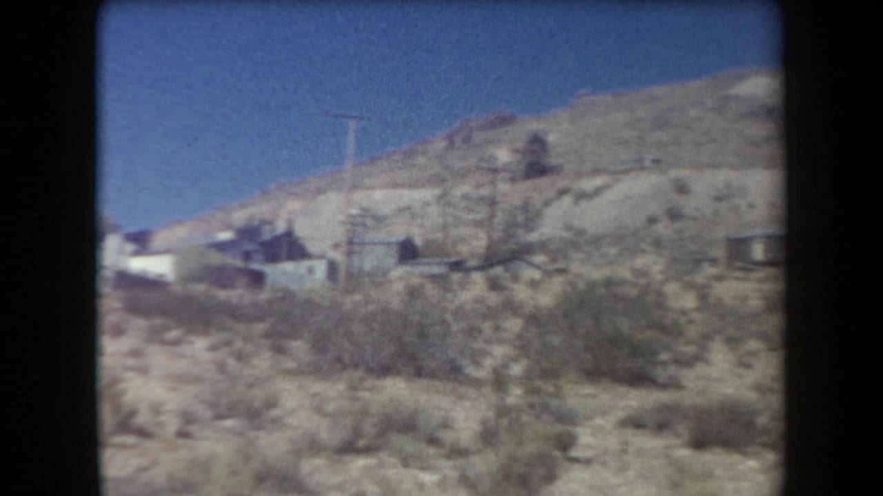 1958 Arizona 8mm frame showing a dry hillside with sparse brush, small houses, and open blue sky.