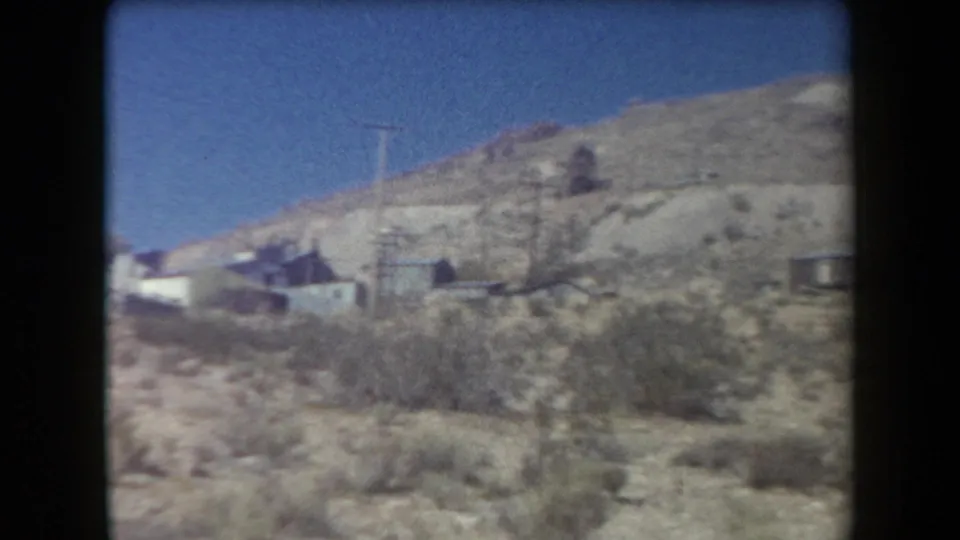 1958 Arizona 8mm frame showing a dry hillside with sparse brush, small houses, and open blue sky.