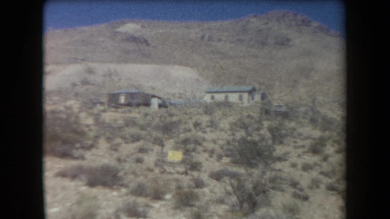 Desert slope with two small houses, scrub brush, and a yellow marker.