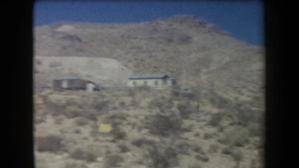 1958 Arizona 8mm view of an arid hillside with scattered brush, small houses, and a yellow marker near the slope.