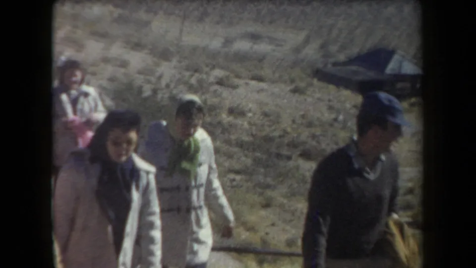 1958 Arizona 8mm frame showing adults walking through scrubby desert terrain below a hillside and small structure.