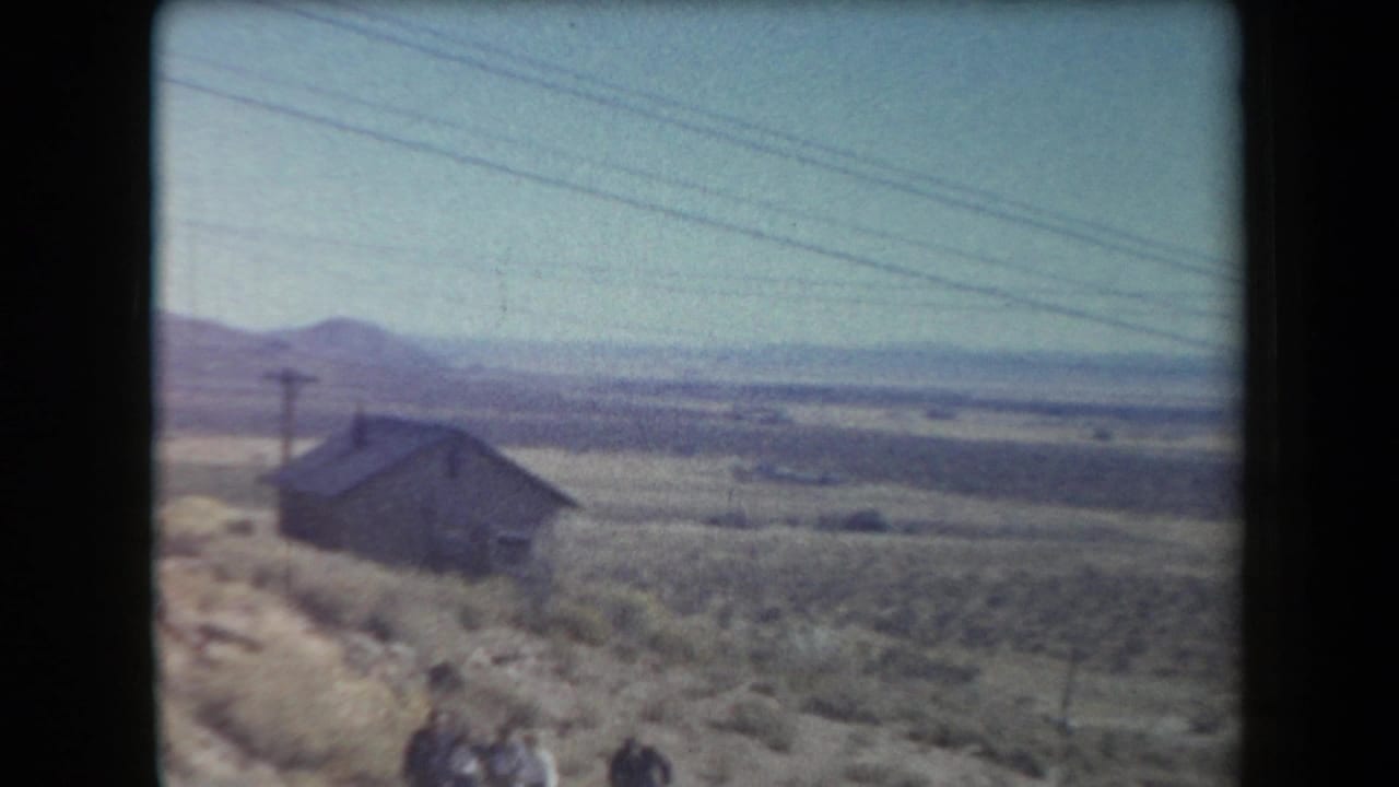 1958 Arizona home-movie frame showing a weathered cabin on desert hillside terrain with utility wires and distant mountains.
