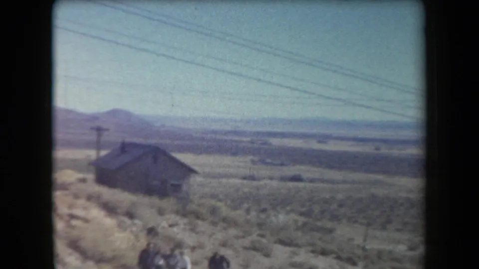 1958 Arizona home-movie frame showing a weathered cabin on desert hillside terrain with utility wires and distant mountains.
