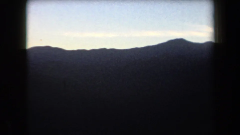 1958 Arizona frame showing a shadowed mountain range with one higher peak rising against a pale blue, lightly clouded sky.