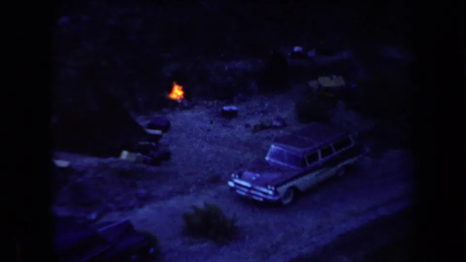 1958 Arizona home-movie frame showing a small orange campfire near a parked station wagon in a dark dirt clearing.