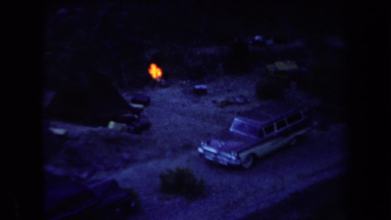 Parked station wagon beside a small campfire in a dark outdoor clearing.