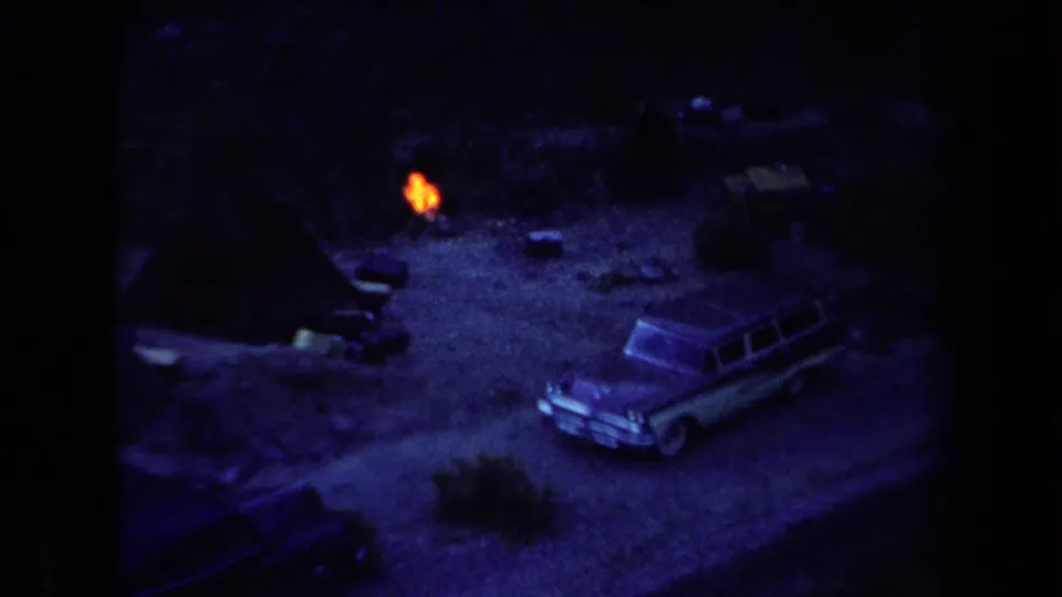 Low-light 1958 Arizona camping footage shows a station wagon parked near a small fire, with dark brush around the clearing.