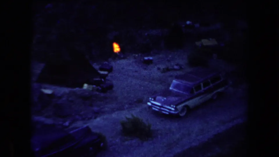 Grainy 8mm frame of a 1958 Arizona campsite, with the station wagon angled beside a small fire in a dim clearing.