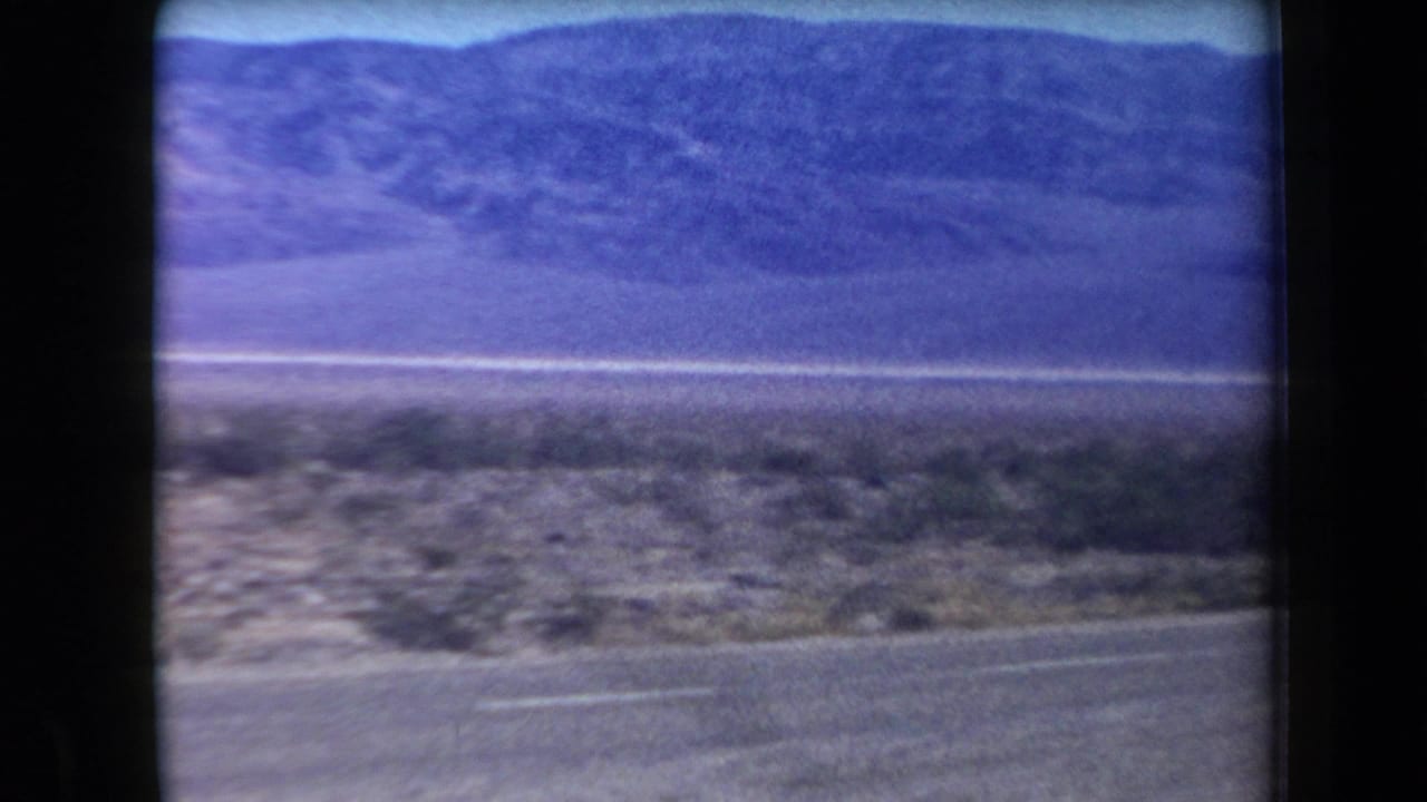1958 Arizona 8mm frame showing a paved roadside, sparse desert brush, flat land, and a distant mountain range.