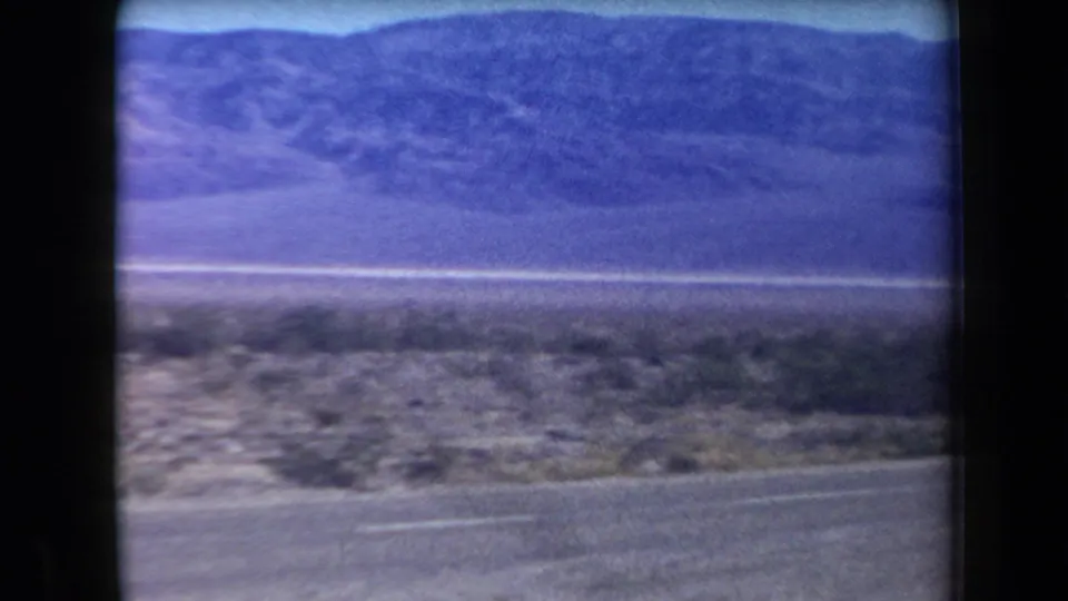 1958 Arizona 8mm frame showing a paved roadside, sparse desert brush, flat land, and a distant mountain range.