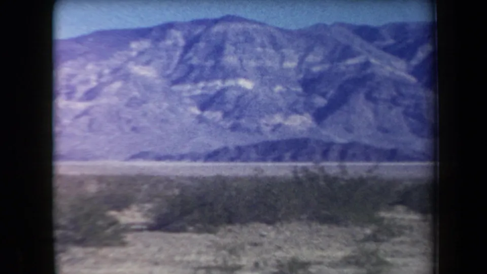 1958 Arizona 8mm still with a wide mountain range, pale desert floor, and low roadside vegetation.