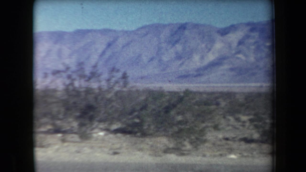 1958 Arizona 8mm frame showing arid roadside shrubs, pale desert ground, and distant mountains under a clear blue sky.