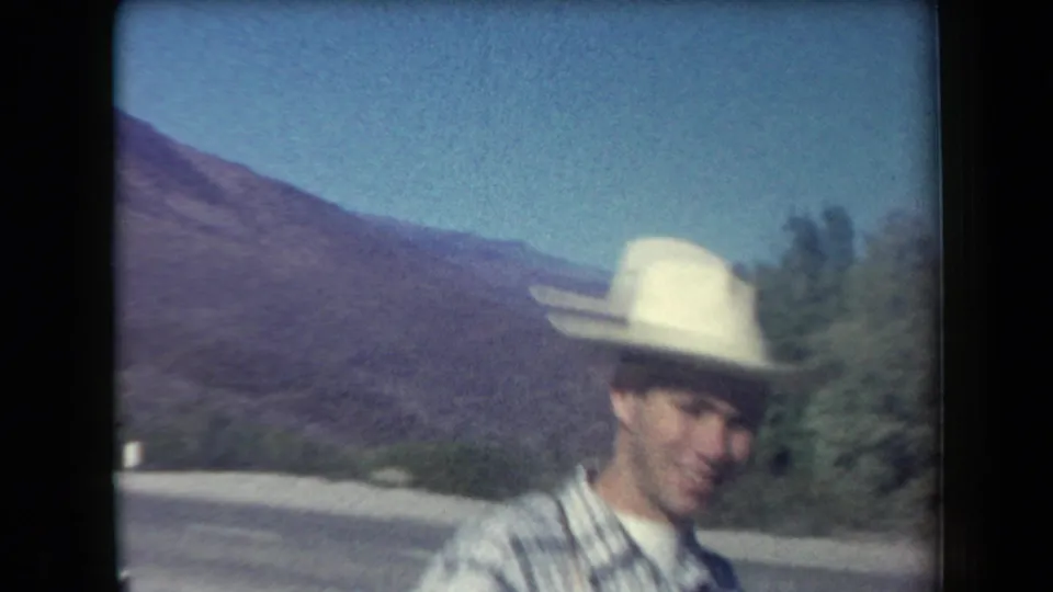 1958 Arizona home-movie frame with a man in a light brimmed hat and plaid shirt beside a road, with desert mountains behind.