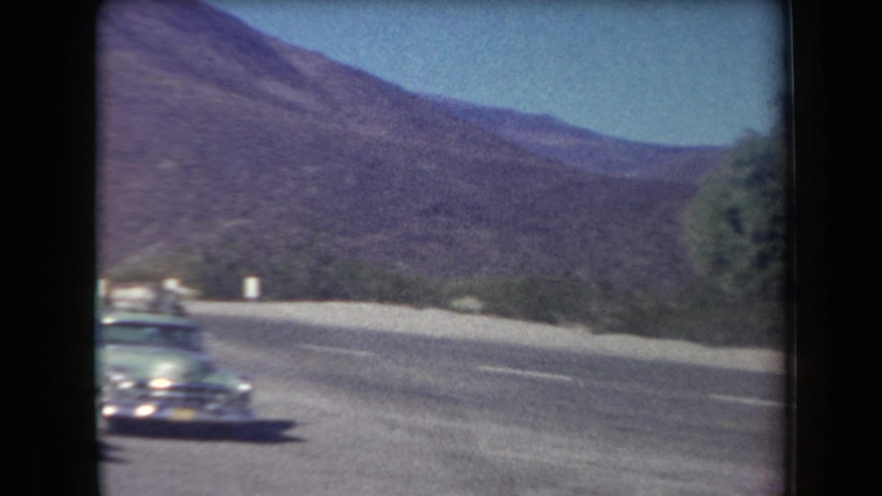 1958 Arizona road scene with a vintage car on a curve and desert mountains beyond, filmed in soft 8mm color.
