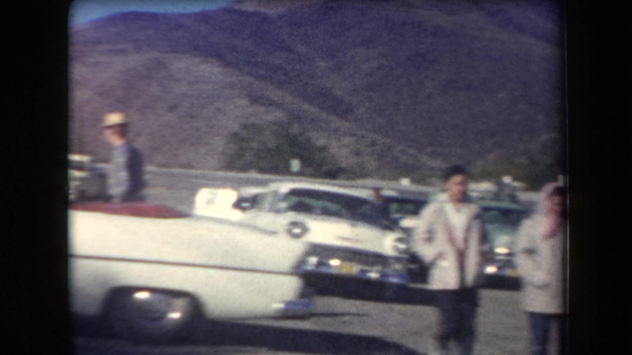 Travelers in period clothing stand beside a white convertible and parked cars near a roadside stop in Arizona.
