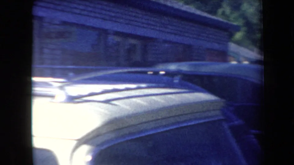 Soft 8mm view of parked 1950s car roofs near a roadside shop or building, with trees and deep shade behind.