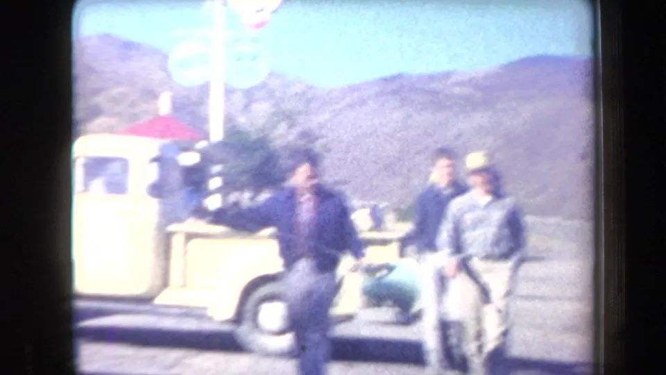 Adult men appear beside a yellow pickup at an Arizona roadside motel area, with dry mountains and a swim-pool sign in the background.