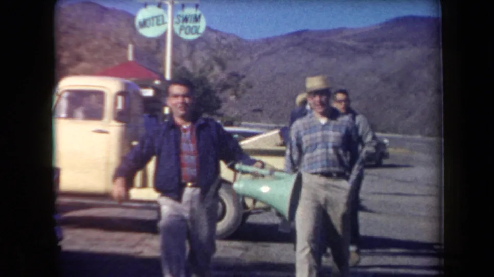 In the 1958 Arizona roadside scene, two adult men approach the camera near a yellow pickup, a green car, and a visible swim-pool sign.
