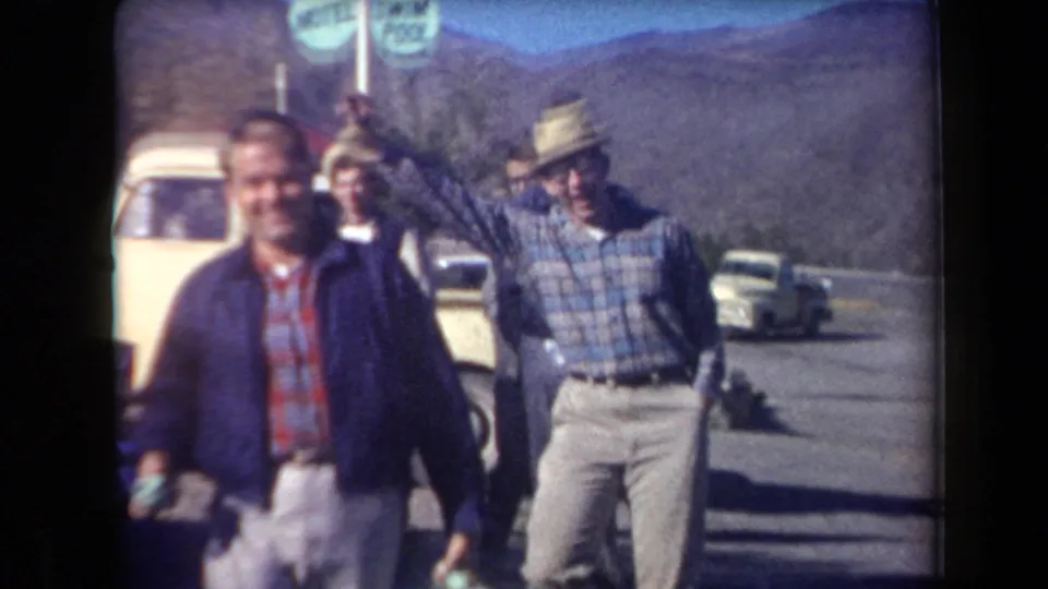 Adult men move toward the camera in Arizona, one carrying a green bullhorn while another raises his fist beside roadside vehicles and mountains.
