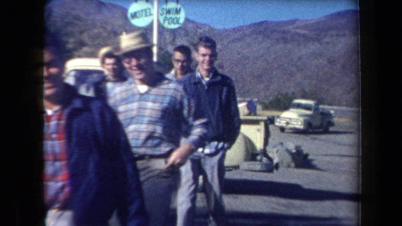 1958 Arizona home-movie frame showing men near a roadside motel area, with a swim pool sign, mountains, and a white pickup truck visible.
