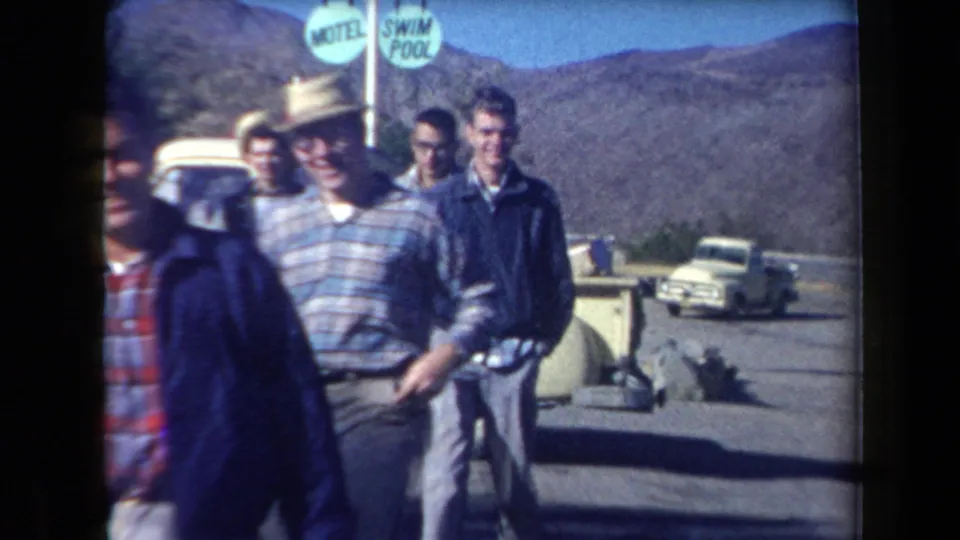 1958 Arizona home-movie frame showing men near a roadside motel area, with a swim pool sign, mountains, and a white pickup truck visible.