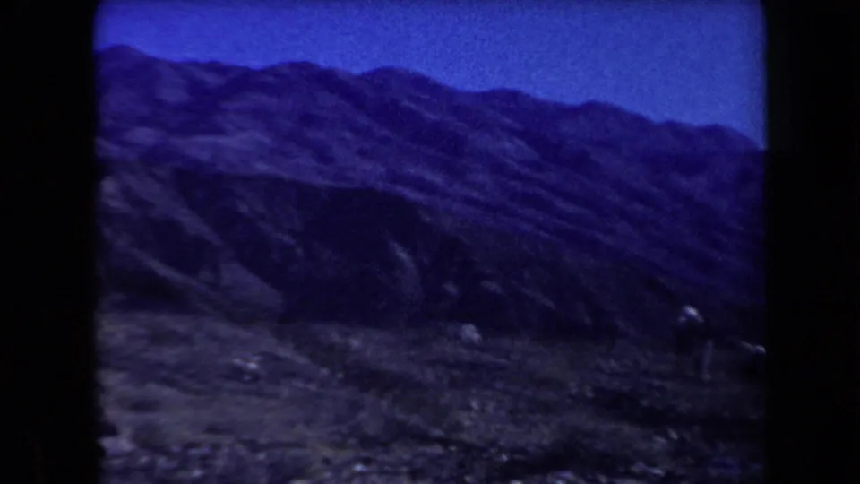 1958 Arizona 8mm frame showing rocky ground and distant mountain ridges with small figures faintly visible in the landscape.