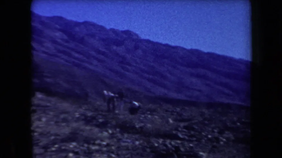 1958 Arizona home-movie frame with distant walkers crossing rocky desert terrain below broad mountain slopes.