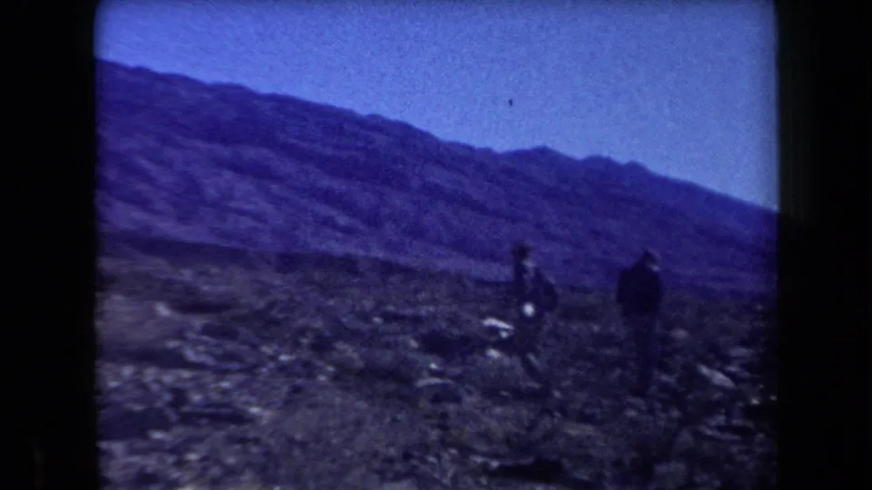 1958 Arizona 8mm frame showing people in dark clothing on a rocky desert slope with mountains stretching across the background.