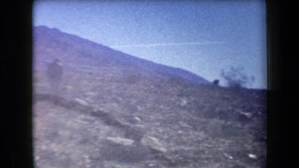 1958 Arizona home-movie frame showing a sunlit rocky hillside, distant mountain slope, and a small figure near the ridge line.