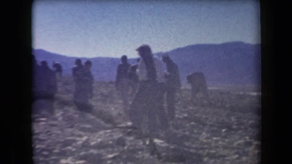 1958 Arizona 8mm frame showing people walking and pausing on rocky terrain with hills in the background.