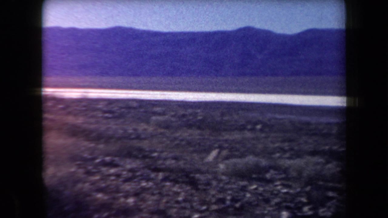1958 Arizona desert view with rocky ground, a pale salt-flat strip, distant mountains, and a blue sky in soft 8mm color.