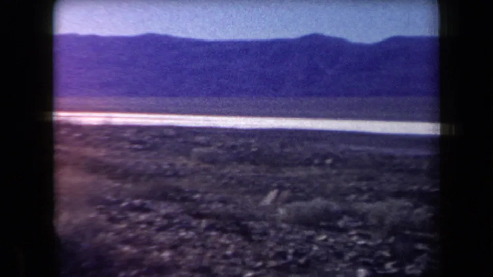 1958 Arizona desert view with rocky ground, a pale salt-flat strip, distant mountains, and a blue sky in soft 8mm color.