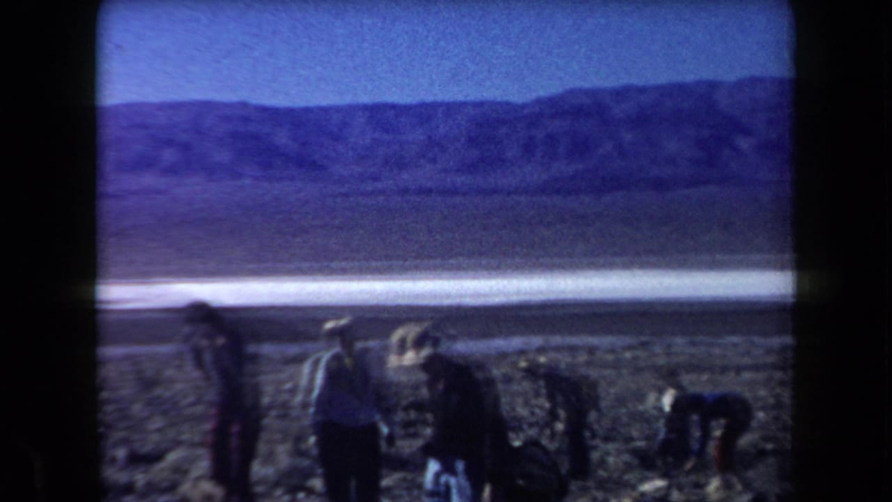 Several people stand and bend on a rocky desert floor with mountains and a pale flat area beyond.